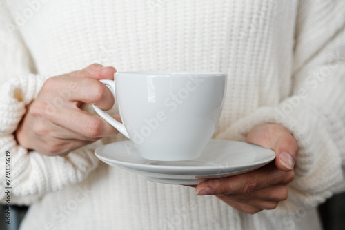 Woman in white knitted sweater holding cup of coffee. Female hands with cup of tea. Close up