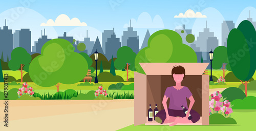 sad man drunk beggar sitting outdoor in cardboard box with bottle of alcohol homeless jobless unemployment concept city urban park cityscape background horizontal flat full length