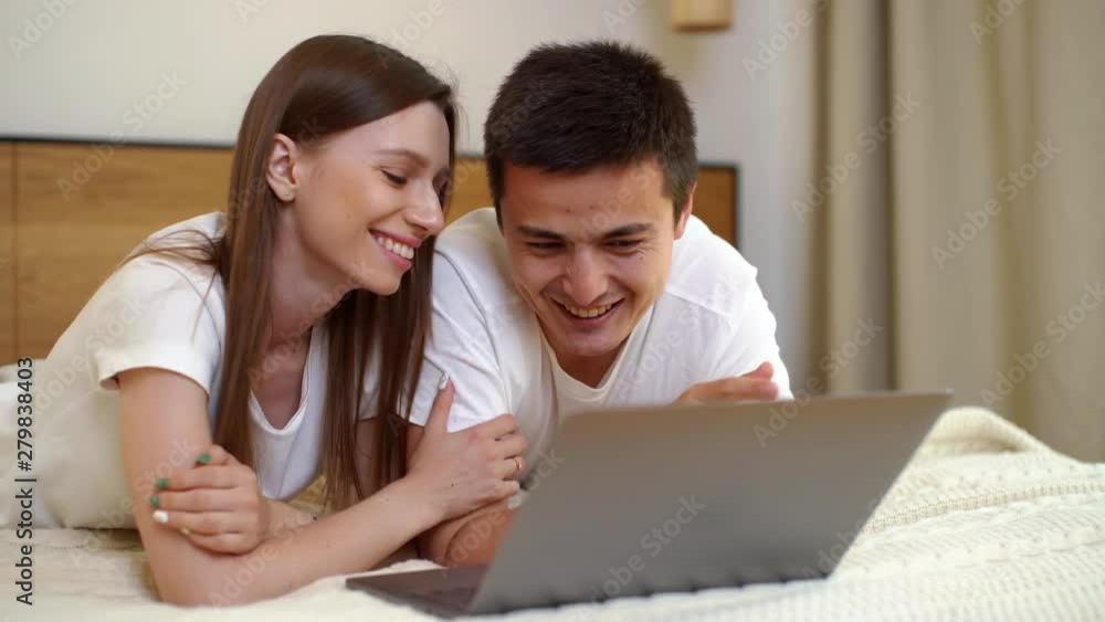 Young happy caucasian young man and woman lying on the bed, talking and using laptop in bright bedroom Young happy caucasian young man and woman lying on the bed, talking and using laptop in bright bedroom