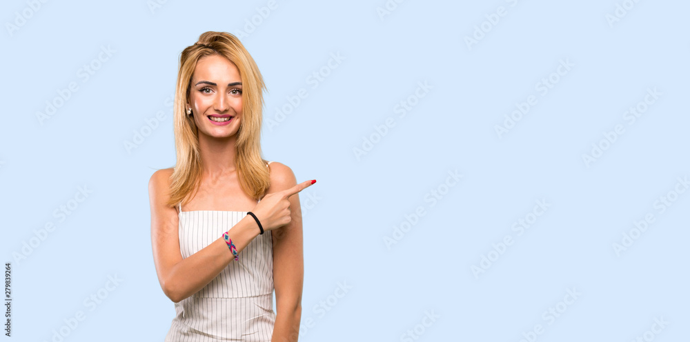 Young blonde woman pointing to the side to present a product over isolated blue background Young blonde woman pointing to the side to present a product over isolated blue background