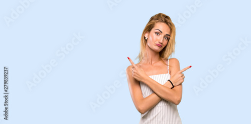 Young blonde woman pointing to the laterals having doubts over isolated blue background
