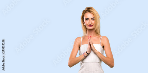 Young blonde woman keeps palm together. Person asks for something over isolated blue background