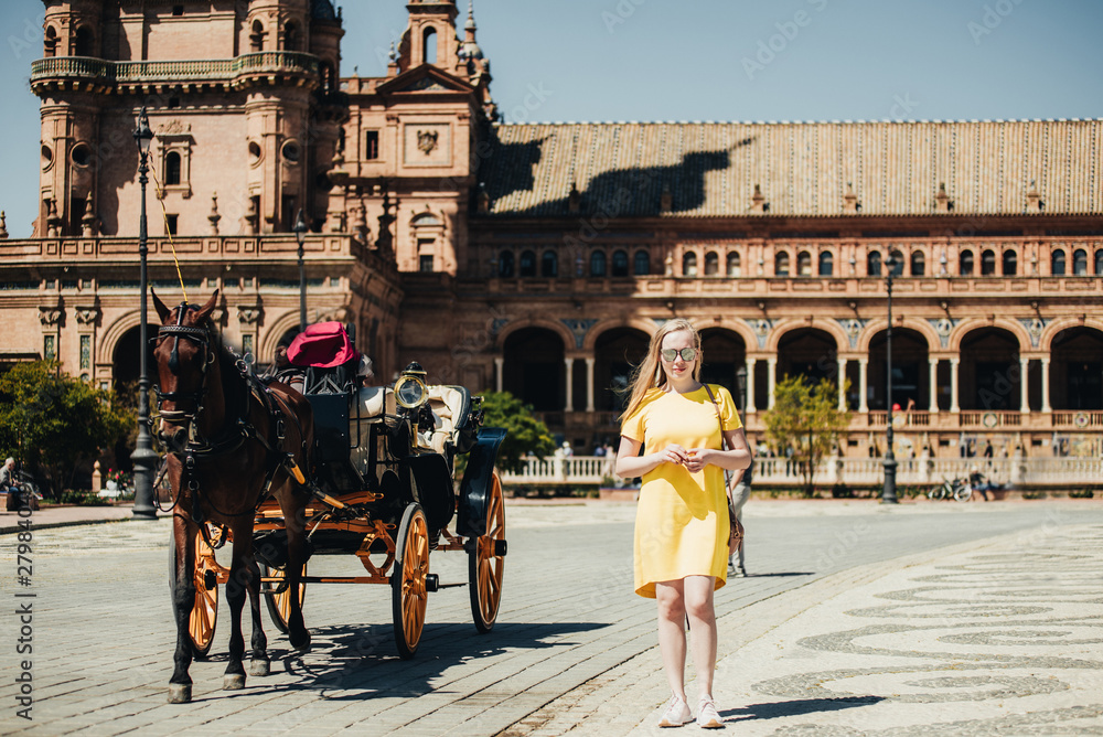 Fototapeta premium Summer outdoor portrait of young beautiful woman in yellow dress. Cheerful blond girl walking between old stone houses in city square. Enjoying vacation in Portugal. Travel concept