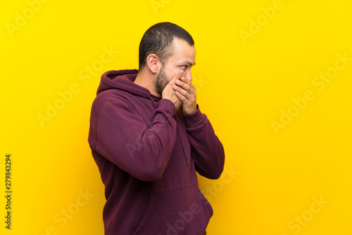 Colombian man with sweatshirt over yellow wall covering mouth and looking to the side © luismolinero