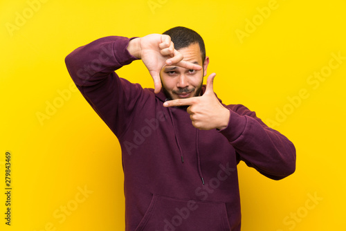 Colombian man with sweatshirt over yellow wall focusing face. Framing symbol