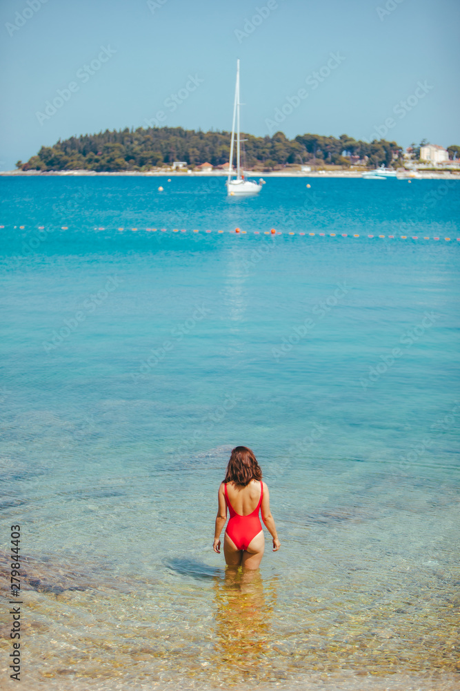 sexy woman in red swimsuit standing in sea water. coastline yacht on background sexy woman in red swimsuit standing in sea water. coastline yacht on background