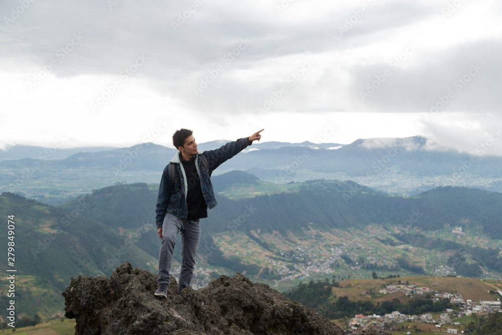 Young Latino on top of a cliff pointing up while smiling with motañas behind him- man exploring Young Latino on top of a cliff pointing up while smiling with motañas behind him- man exploring