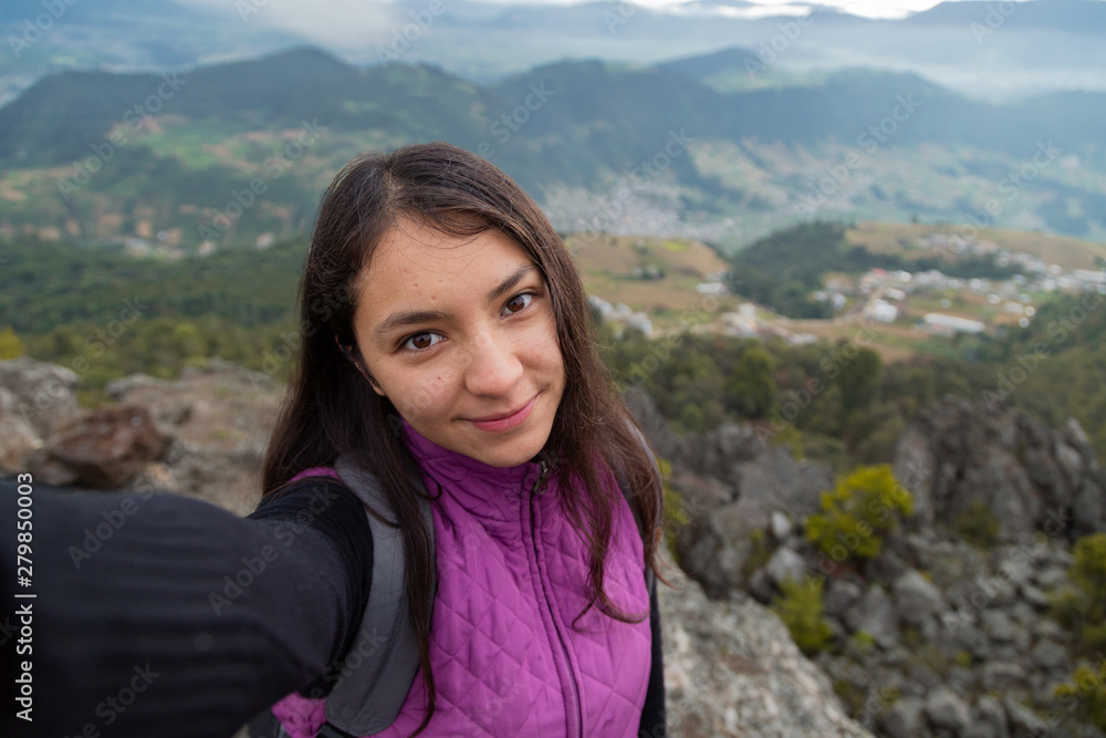 Beautiful latin woman taking a selfie on top of the mountain with the small town behind her- Hispanic traveler