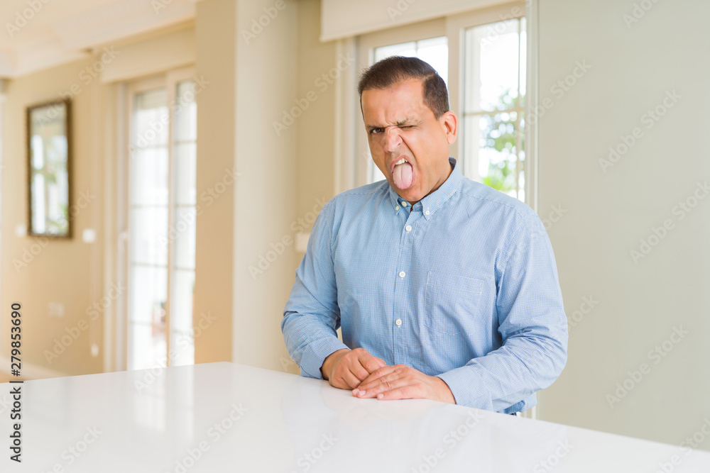 Middle age man sitting at home sticking tongue out happy with funny expression. Emotion concept.
