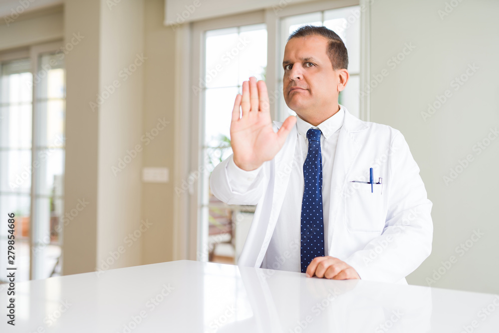 Fototapeta premium Middle age doctor man wearing medical coat at the clinic doing stop sing with palm of the hand. Warning expression with negative and serious gesture on the face.