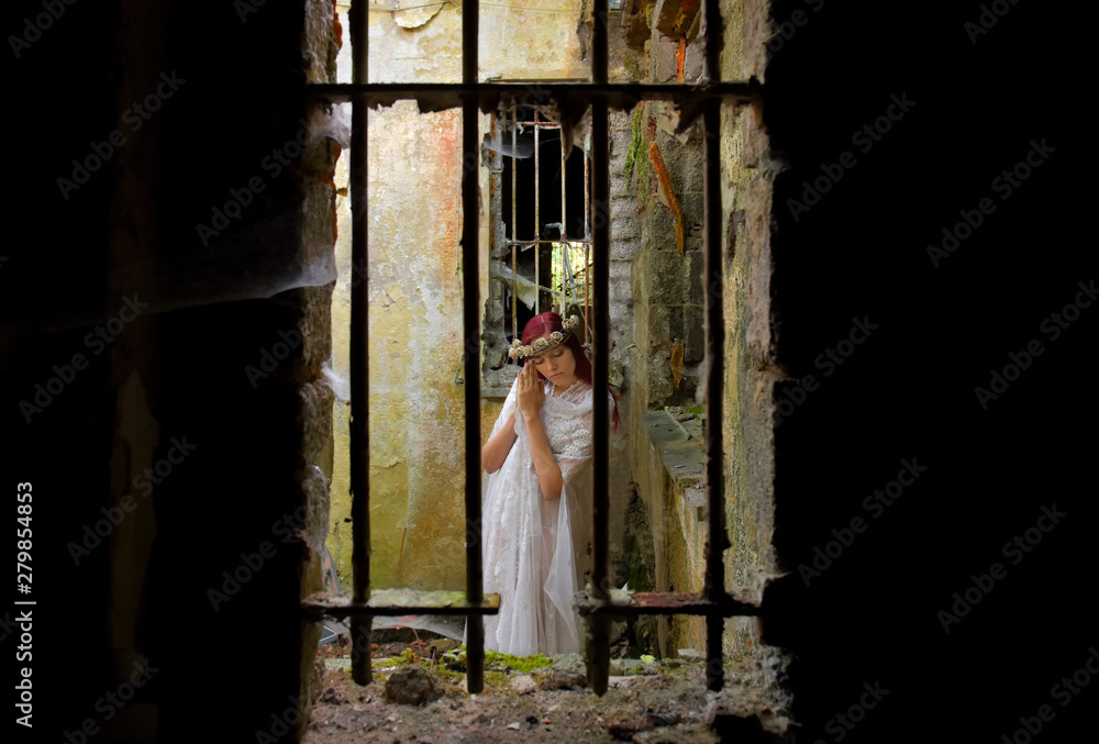 A young woman prays inside a jail ruin. She is  dressed in white and wears a thorn wreath on  her head. 