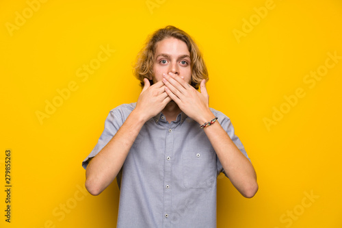 Blonde man over isolated yellow background covering mouth with hands