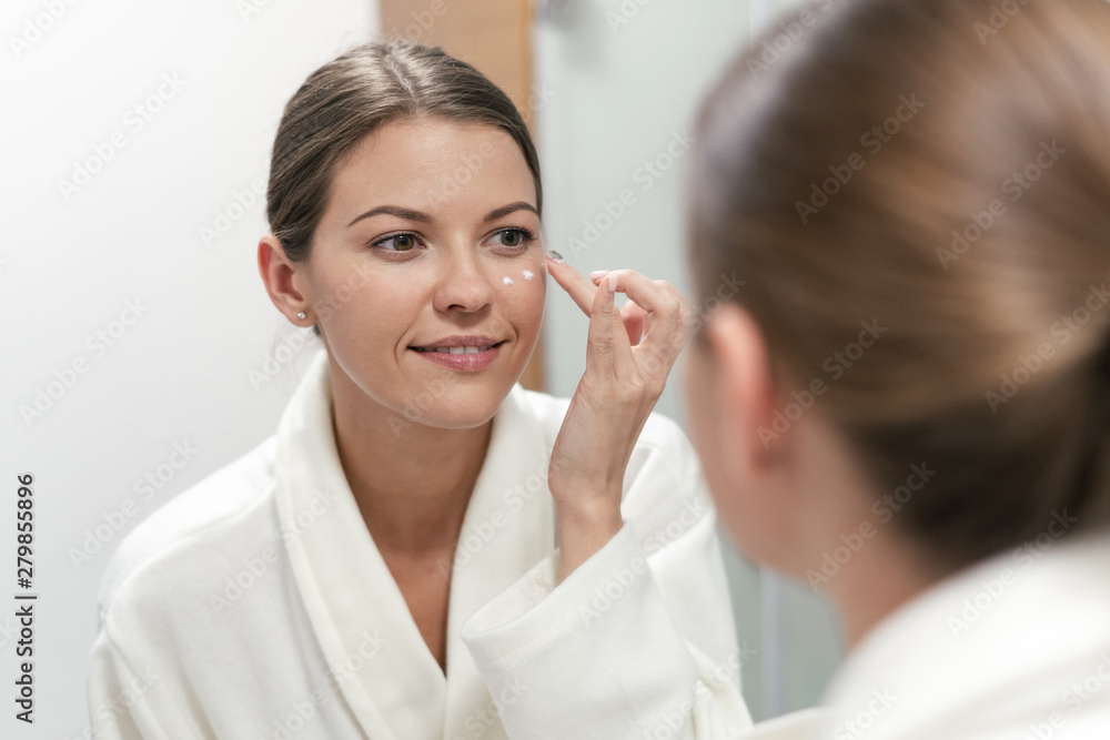 Woman standing in bathroom, applying cream on face Woman standing in bathroom, applying cream on face