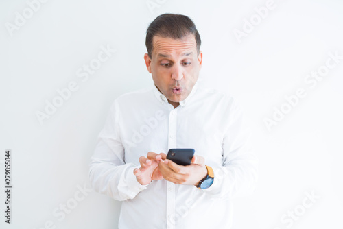 Middle age man using smartphone over white wall scared in shock with a surprise face, afraid and excited with fear expression © Krakenimages.com