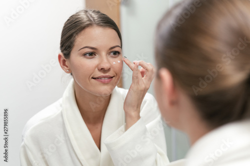 Woman standing in bathroom, applying cream on face