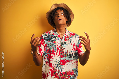 Afro american man with dreadlocks wearing floral shirt and hat over isolated yellow background amazed and surprised looking up and pointing with fingers and raised arms.