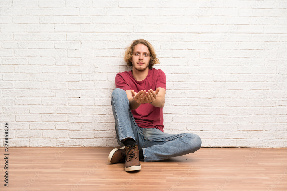 Blonde man sitting on the floor holding copyspace imaginary on the palm to insert an ad Blonde man sitting on the floor holding copyspace imaginary on the palm to insert an ad