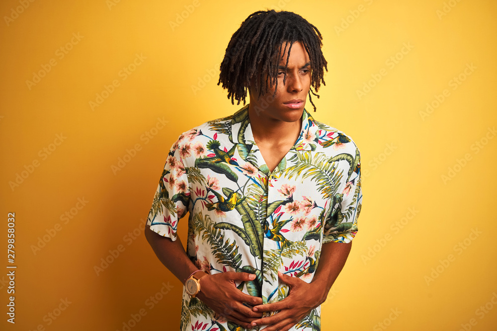 Afro man with dreadlocks on vacation wearing summer shirt over isolated yellow background with hand on stomach because nausea, painful disease feeling unwell. Ache concept.
