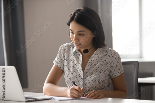 Asian businesswoman wear headset watching webinar make call write notes