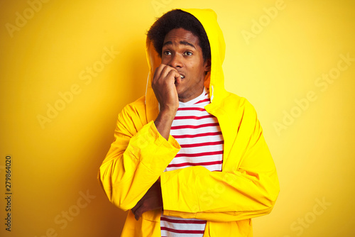 Fotomural African man with afro hair wearing rain coat with hood standing over isolated yellow background looking stressed and nervous with hands on mouth biting nails