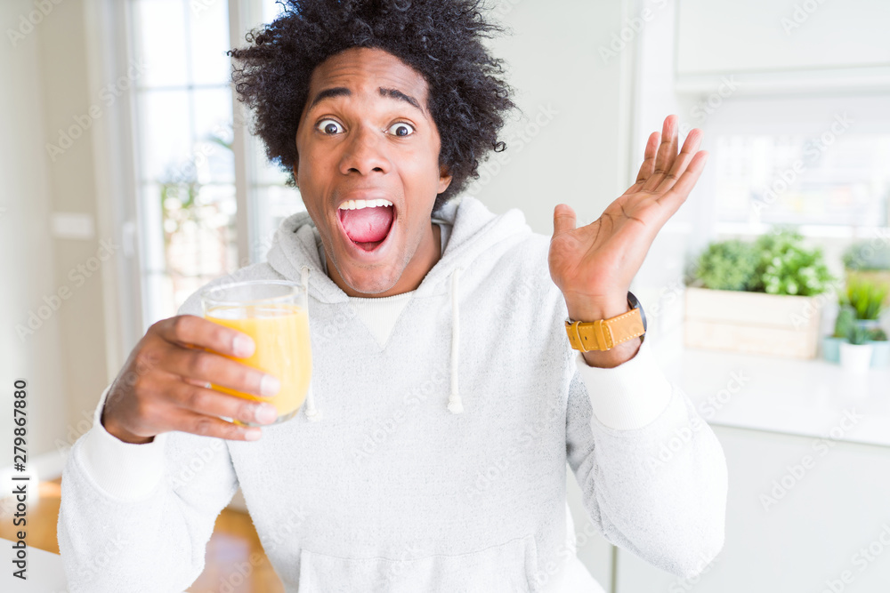 African American man holding and drinking glass of orange juice very happy and excited, winner expression celebrating victory screaming with big smile and raised hands African American man holding and drinking glass of orange juice very happy and excited, winner expression celebrating victory screaming with big smile and raised hands