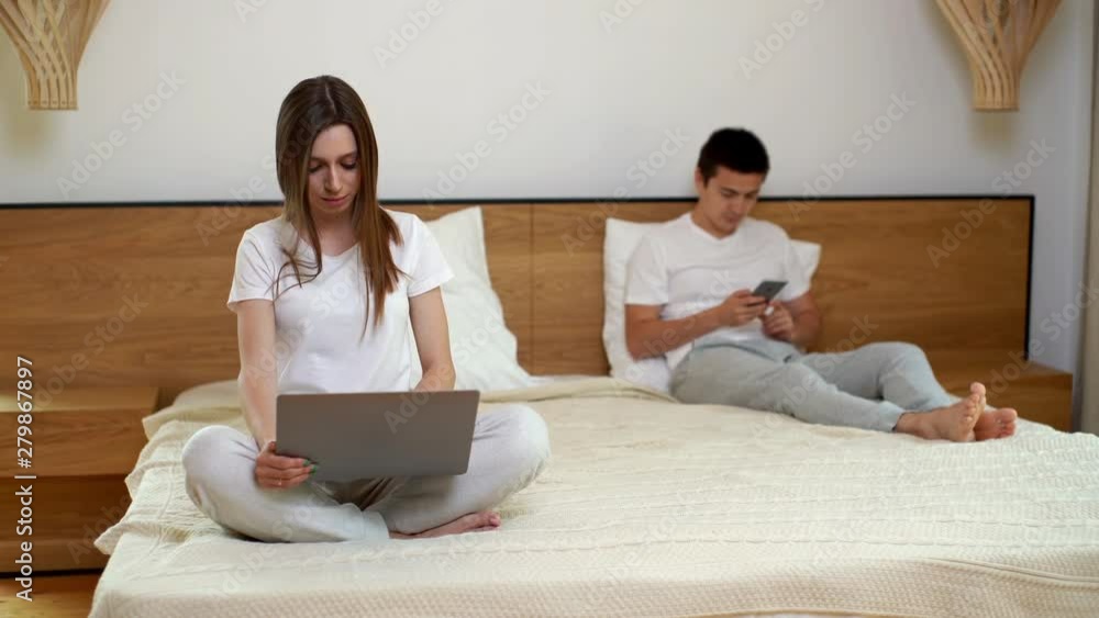 Girl working on laptop sitting in bed. Guy uses phone behind his girlfriend. Man and a woman using mobile phone and laptop while sitting in bed. Girl is typing on notepad, the guy is surfing Internet Girl working on laptop sitting in bed. Guy uses phone behind his girlfriend. Man and a woman using mobile phone and laptop while sitting in bed. Girl is typing on notepad, the guy is surfing Internet