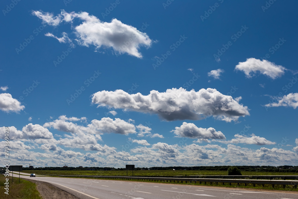 Cumulus clouds in the blue sky over a field with a highway. Sky pattern.