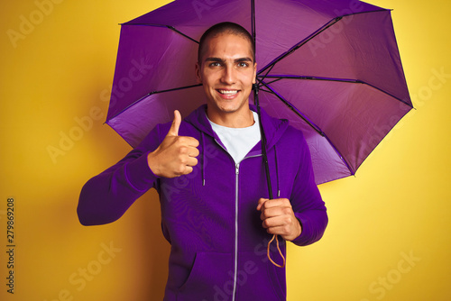 Young handsome man wearing purple umbrella over yellow isolated background happy with big smile doing ok sign, thumb up with fingers, excellent sign