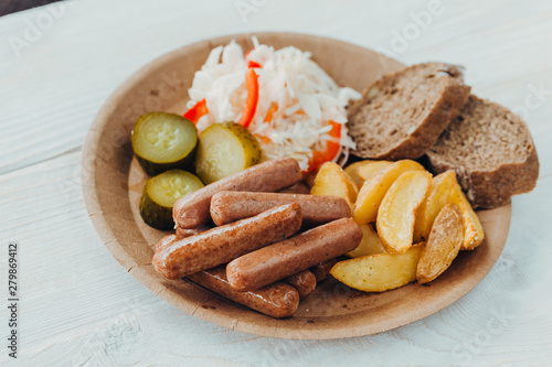 Tasty sausages with fried potatoes  cucumber  cabbage and chlobe in a paper plate on a wooden table.