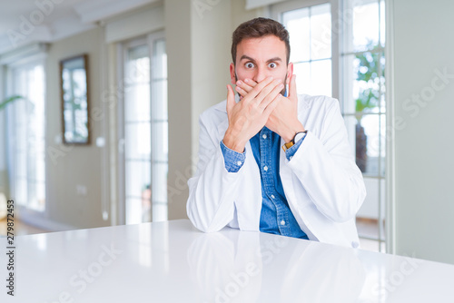 Handsome doctor man wearing medical coat at the clinic shocked covering mouth with hands for mistake. Secret concept.