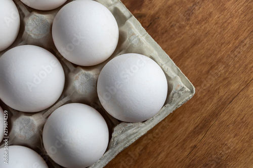 Chicken eggs are raw in the tray. The shell is white in eggs. In one section is an egg shell. On one egg lies a feather of white color. Wooden background. View from above. Close-up.