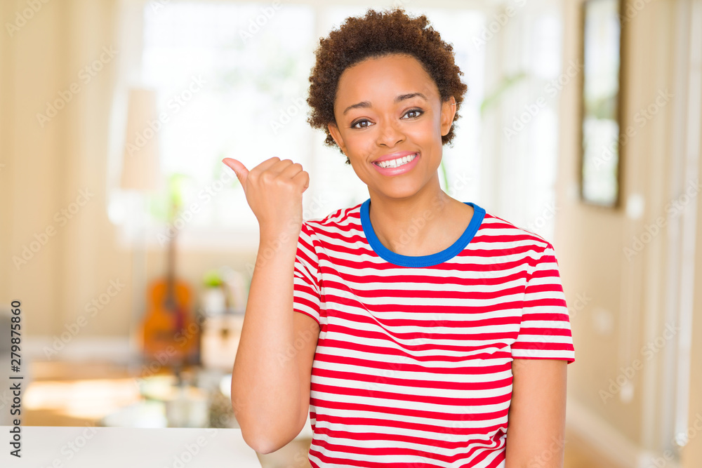 Young beautiful african american woman at home smiling with happy face looking and pointing to the side with thumb up.