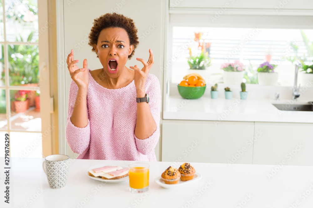 Young african american woman eating breaksfast in the morning at home crazy and mad shouting and yelling with aggressive expression and arms raised. Frustration concept. Young african american woman eating breaksfast in the morning at home crazy and mad shouting and yelling with aggressive expression and arms raised. Frustration concept.