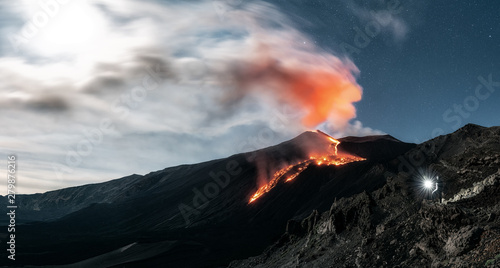 Panoramic image of a Volcano Etna during an incredible eruption at night with full moon. A man lightning up the landscape with a torch