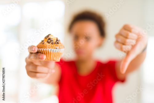 Young african american woman eating chocolate chips muffins with angry face, negative sign showing dislike with thumbs down, rejection concept