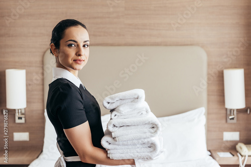 smiling maid holding pile of folded towels near bed and looking at camera in hotel room