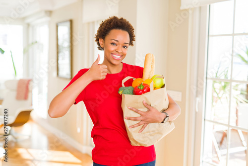 Young african american woman holding paper bag full of fresh groceries happy with big smile doing ok sign, thumb up with fingers, excellent sign