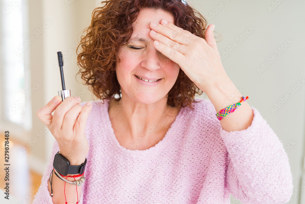 Obraz premium Senior woman putting mascara on eyelashes stressed with hand on head, shocked with shame and surprise face, angry and frustrated. Fear and upset for mistake.