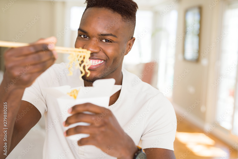 Handsome african man eating asian noodles in a delivery box, smiling enjoying lunch using chopsticks