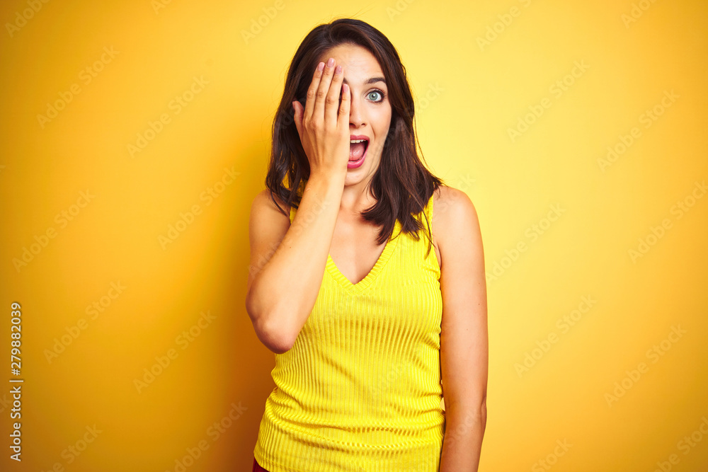 Young beautiful woman wearing t-shirt standing over yellow isolated background covering one eye with hand, confident smile on face and surprise emotion. Young beautiful woman wearing t-shirt standing over yellow isolated background covering one eye with hand, confident smile on face and surprise emotion.