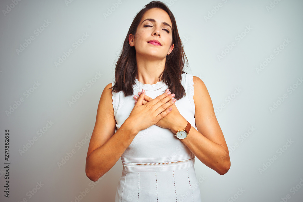 Young beautiful woman wearing dress standing over white isolated background smiling with hands on chest with closed eyes and grateful gesture on face. Health concept. Young beautiful woman wearing dress standing over white isolated background smiling with hands on chest with closed eyes and grateful gesture on face. Health concept.