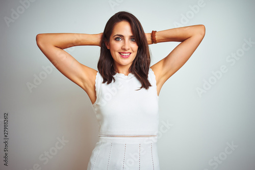 Young beautiful woman wearing dress standing over white isolated background relaxing and stretching, arms and hands behind head and neck smiling happy