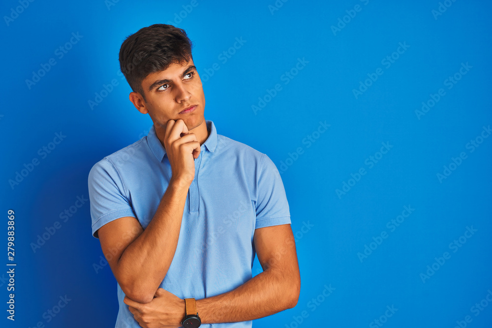 Young indian man wearing casual polo standing over isolated blue background with hand on chin thinking about question, pensive expression. Smiling with thoughtful face. Doubt concept. Young indian man wearing casual polo standing over isolated blue background with hand on chin thinking about question, pensive expression. Smiling with thoughtful face. Doubt concept.