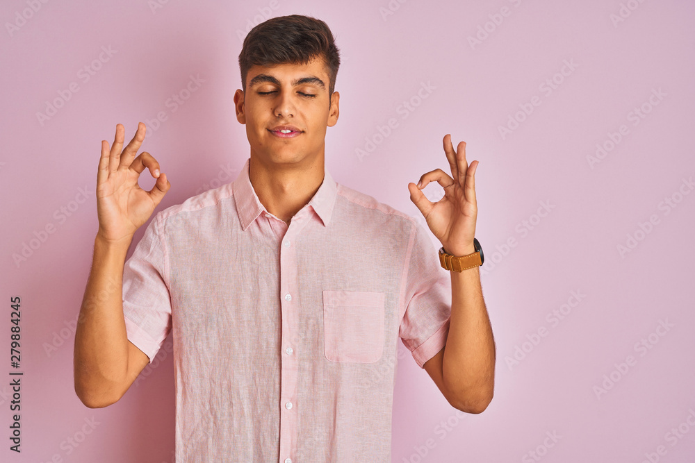 Young indian man wearing casual shirt standing over isolated pink background relax and smiling with eyes closed doing meditation gesture with fingers. Yoga concept. Young indian man wearing casual shirt standing over isolated pink background relax and smiling with eyes closed doing meditation gesture with fingers. Yoga concept.