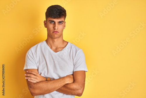Young indian man wearing white t-shirt standing over isolated yellow background skeptic and nervous, disapproving expression on face with crossed arms. Negative person.