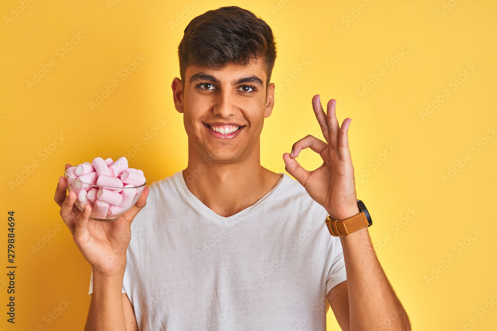 Young indian man holding bowl with marshmallows over isolated yellow background doing ok sign with fingers, excellent symbol Young indian man holding bowl with marshmallows over isolated yellow background doing ok sign with fingers, excellent symbol