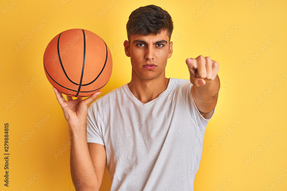 Young indian sportsman holding basketball ball standing over isolated yellow background pointing with finger to the camera and to you, hand sign, positive and confident gesture from the front Young indian sportsman holding basketball ball standing over isolated yellow background pointing with finger to the camera and to you, hand sign, positive and confident gesture from the front