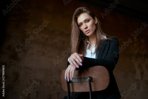 Studio portrait of young beautiful sensual woman in blak suit sitting on wooden chair against dark background.
