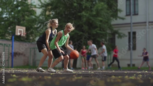 Tracking shot of two college girls playing basketball on outdoor court in slow motion. Female friends practicing defender and forward roles, group of students exercising in background