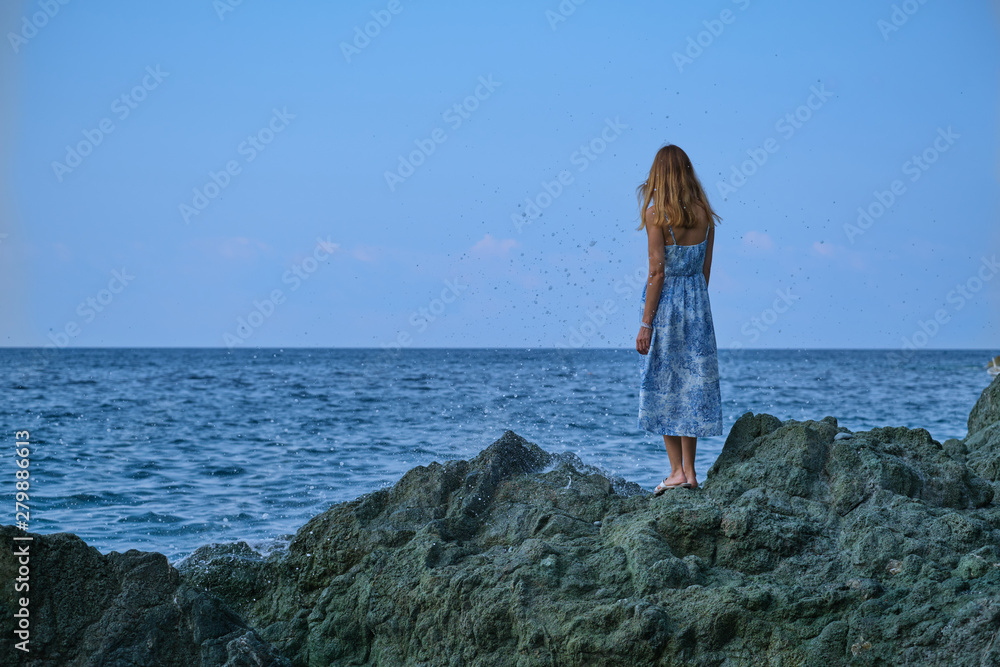 A beautiful young girl in a blue dress is standing on a rock and looking at the sea.